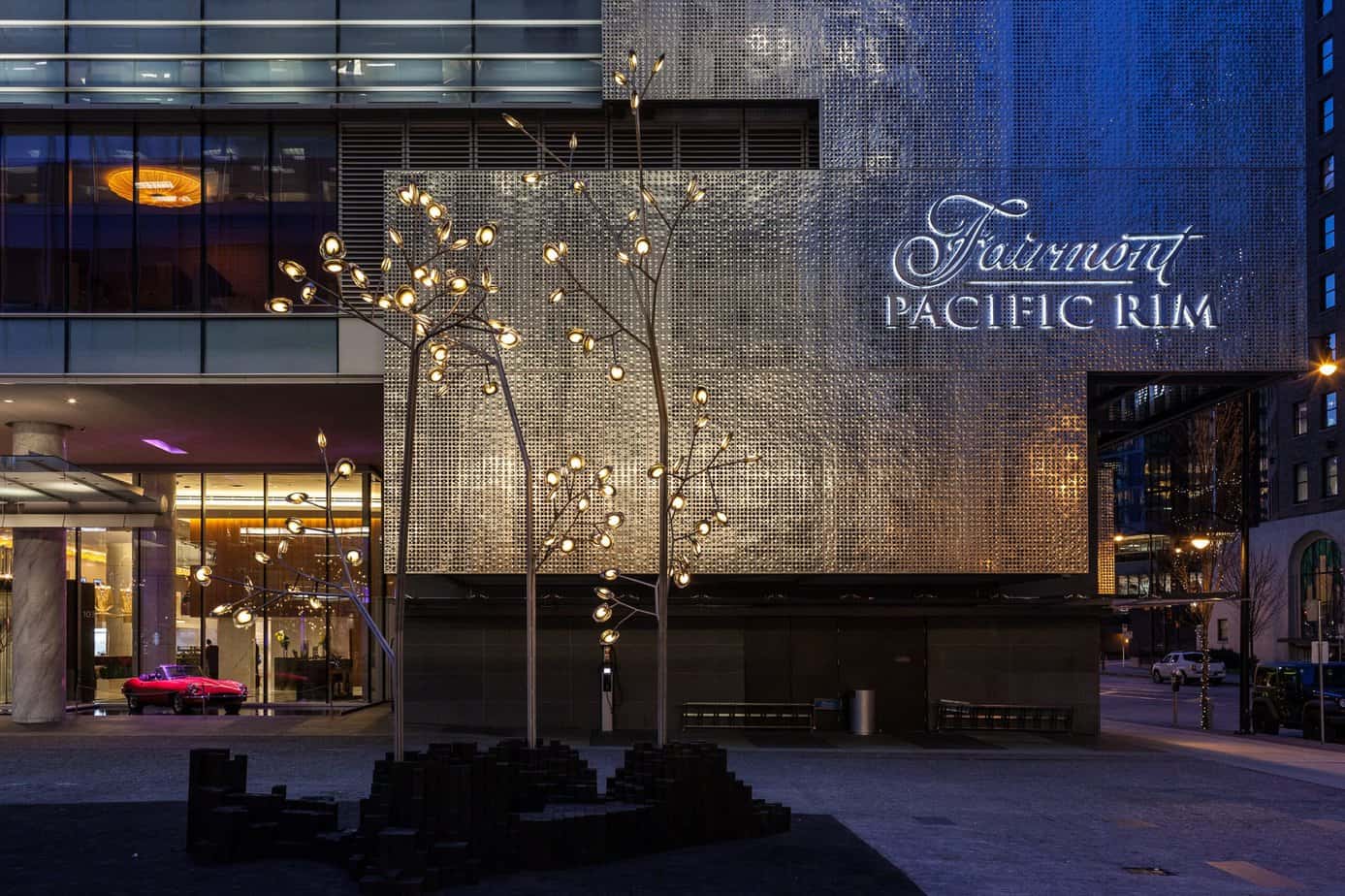 Front facing of the hotel with a silvery background with "Fairmont Pacific Rim" written on front. "Fairmont" is in cursive. Fake tree lights in the middle with a red vintage looking car in the window. Fairmont Pacific Rim is one of the best Vancouver hotels with outdoor pools.