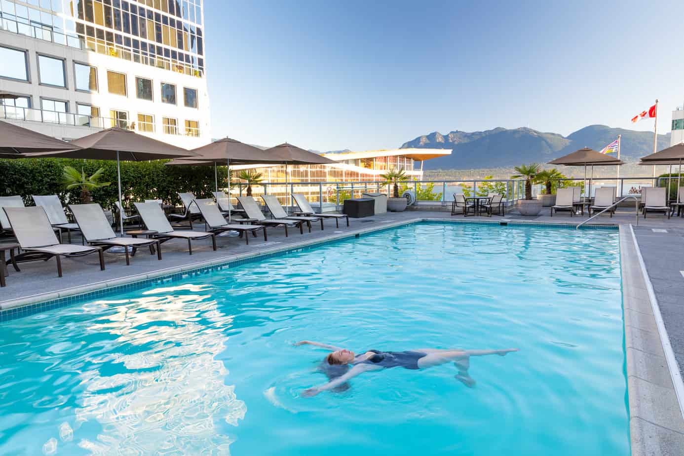 A women floating on her back with her arms up relaxing in the outdoor roof top pool. There is a view of the mountains and Northshore and part of the Vancouver convention Centre building. There are many lounge chairs surrounding the pool.