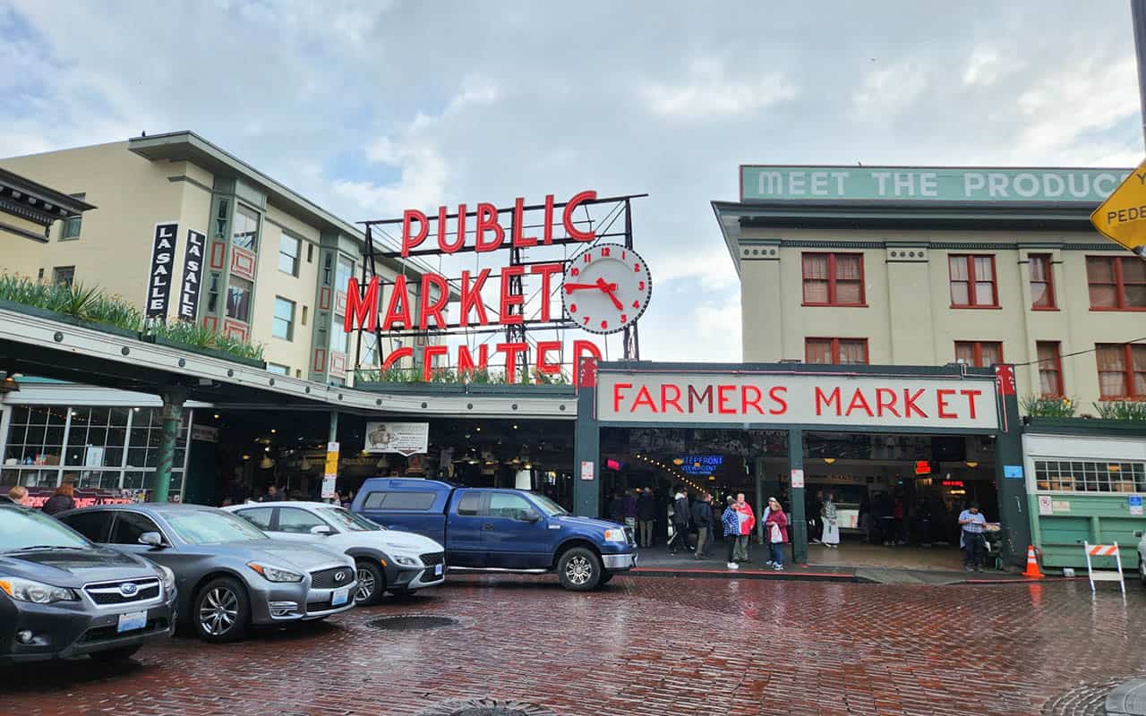 Cobblestone streer with buildings in a greenish yellow colour with a large red neon words that says "Public Market Center" and it has a clock on it beside the word market. Then below it is says "Farmers Market" in the same red neon. And four cars parked on the left side and people populating inside the market. Pike Place Public Market is one of the tourist attractions in Seattle.