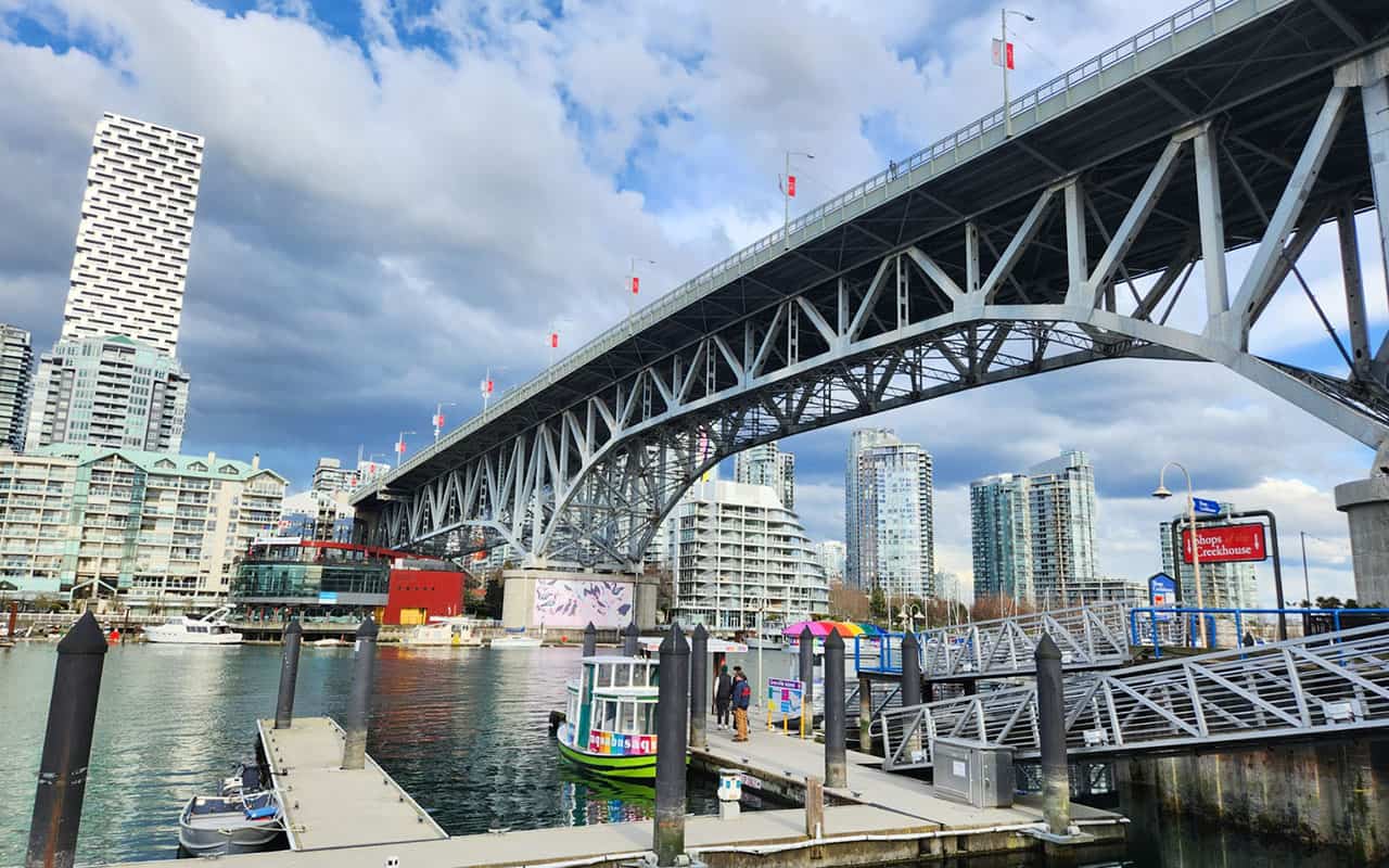 Bottom angle of the Granville Island Bridge on the right with a Vancouver Aquabus with people waiting on the small dock and buildings along the waterfront.