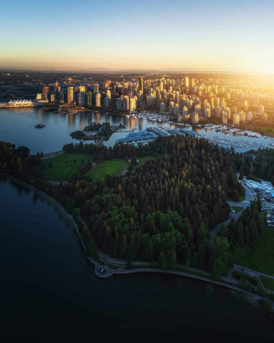Birds eye view during either sunset or sunrise of the green Stanley Park below and the city of Vnacouver and buildings in the distance and continues on. Stanley Park is one of the most beloved urban parks in Vancouver British Columbia.