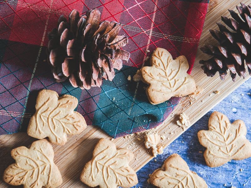 Maple leaf shaped cookies on a cutting board with pinecones and a red and green plaid cloth underneath the cookies and pinecones on a blue table.