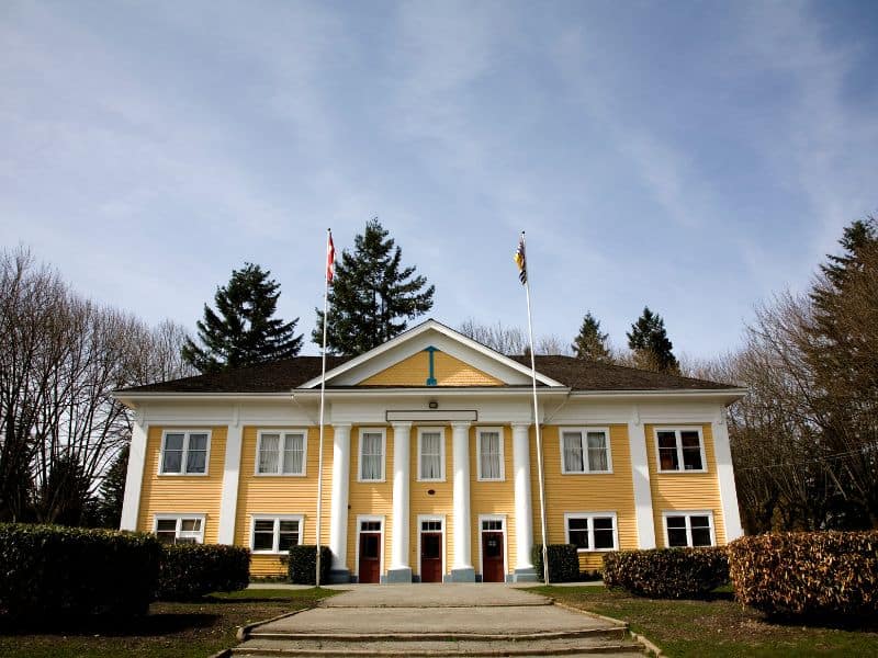 A yellow old fashion building with four white columns on the building. With a Canada flag on the left and a British Columbia flag on the right. The yellow Langley Town Hall is well known for the backdrop of many tv shows and movies.